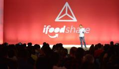 Um palestrante no palco de um evento corporativo falando para uma plateia lotada, com um grande painel vermelho ao fundo exibindo o logotipo 