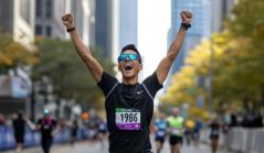 Corredor de maratona comemorando com os braços erguidos e expressão de euforia ao cruzar a linha de chegada em uma avenida cercada por prédios e outros competidores.