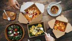 Vista superior de uma mesa de madeira com diversas embalagens de papel abertas contendo pratos variados como massas, risoto de camarão, salada e quiche, com mãos utilizando garfos para comer.