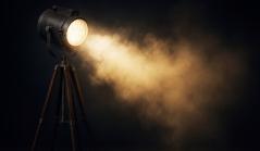 Um refletor de estilo vintage montado em um tripé de madeira, projetando um feixe de luz amarelada através de uma névoa ou fumaça em um ambiente escuro.