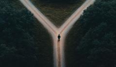 Vista aérea de uma pessoa solitária parada em uma bifurcação de uma estrada de terra, cercada por árvores densas, simbolizando uma escolha ou tomada de decisão.