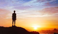 Silhueta de uma pessoa no topo de uma rocha contemplando o pôr do sol sobre o mar, transmitindo reflexão e visão de futuro.