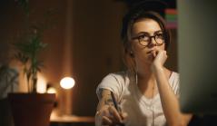 Mulher jovem de óculos e boné, com expressão pensativa, usando uma mesa digitalizadora em frente ao computador em um ambiente com iluminação quente.