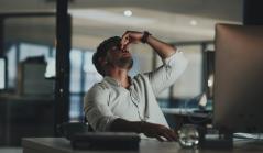 Homem sentado em um escritório escuro aperta a ponta do nariz com os olhos fechados, demonstrando cansaço ou estresse diante do computador.