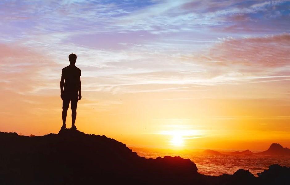 Silhueta de uma pessoa no topo de uma rocha contemplando o pôr do sol sobre o mar, transmitindo reflexão e visão de futuro.