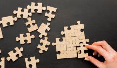 Vista superior de uma mão encaixando a última peça de um quebra-cabeça de madeira sobre uma superfície preta, com várias outras peças espalhadas ao lado.