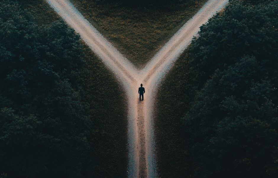 Vista aérea de uma pessoa solitária parada em uma bifurcação de uma estrada de terra, cercada por árvores densas, simbolizando uma escolha ou tomada de decisão.