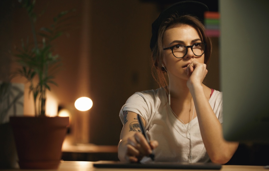 Mulher jovem de óculos e boné, com expressão pensativa, usando uma mesa digitalizadora em frente ao computador em um ambiente com iluminação quente.