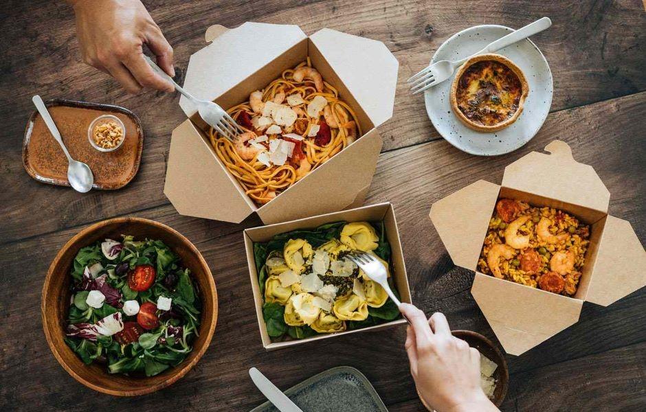 Vista superior de uma mesa de madeira com diversas embalagens de papel abertas contendo pratos variados como massas, risoto de camarão, salada e quiche, com mãos utilizando garfos para comer.