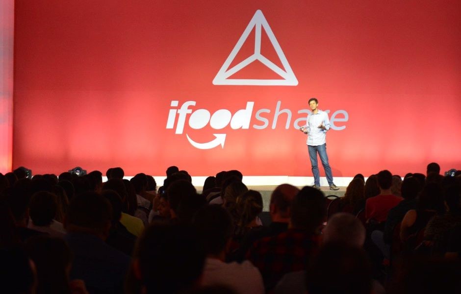 Um palestrante no palco de um evento corporativo falando para uma plateia lotada, com um grande painel vermelho ao fundo exibindo o logotipo 